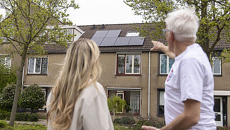 Een energiecoach en en vrouw staan samen voor een huis met zonnepanelen op het dak. Man wijst naar de zonnepanelen. 